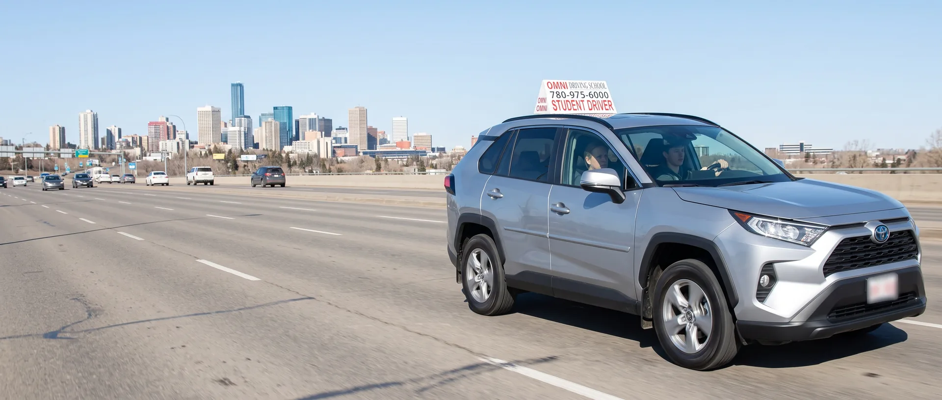 The Omni Driving School car on a wide city road, the downtown skyline behind