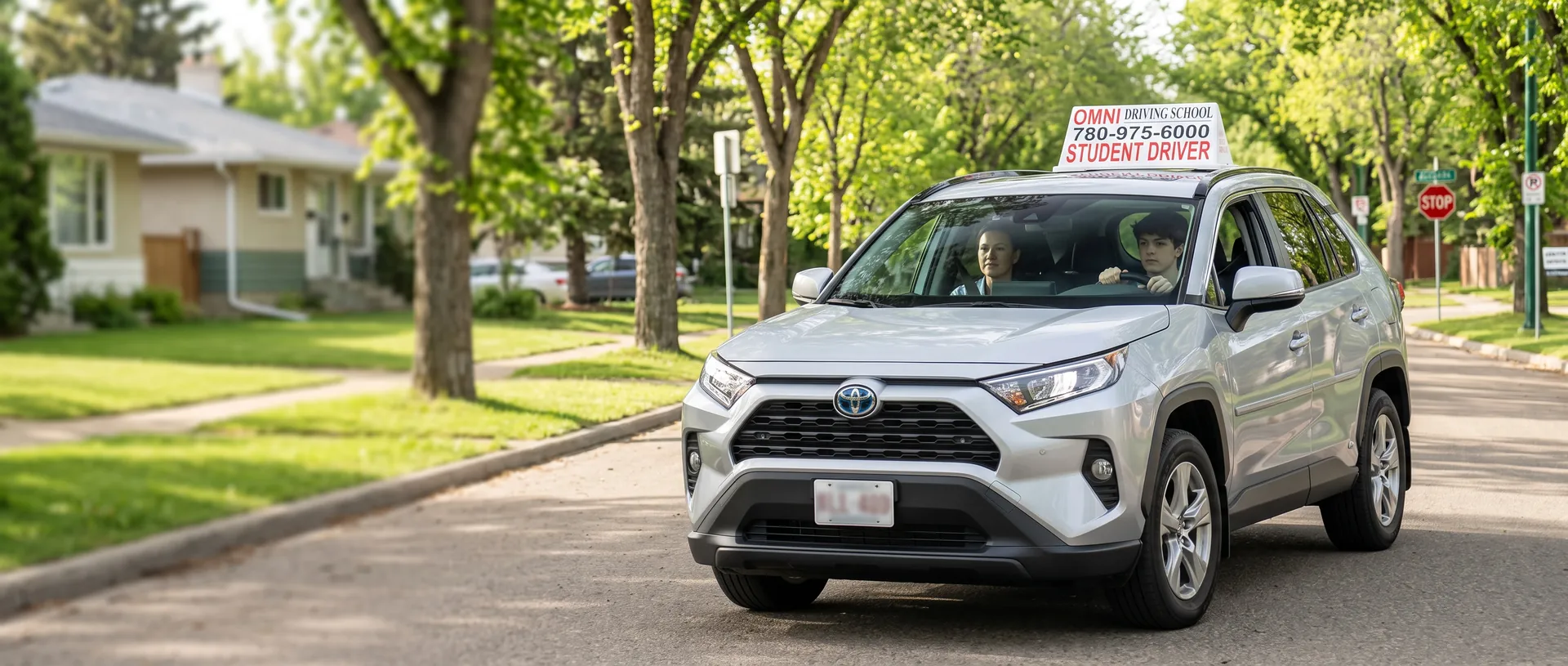 A student drives the Omni Driving School car on a leafy street, the instructor beside